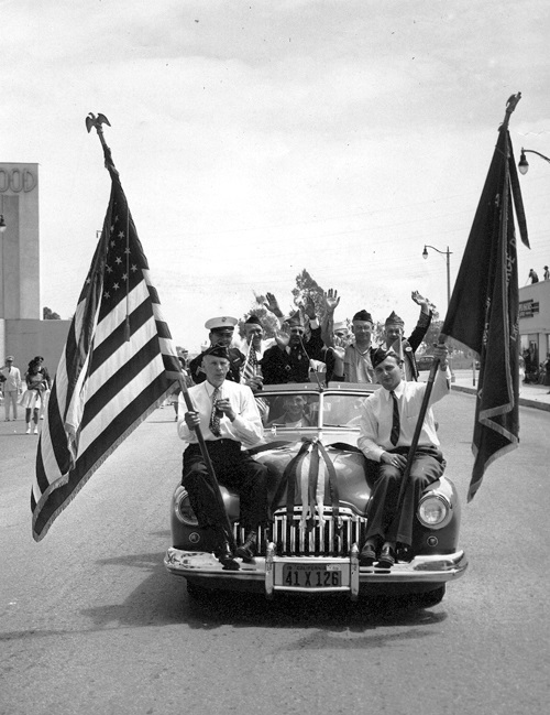 Men ride with flags on the hood of a car in Lakewood Village in 1949